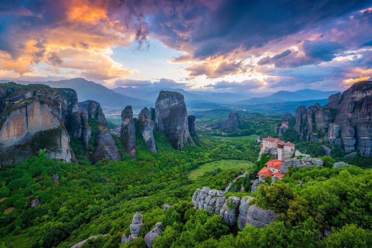 Scenic view of Meteora, Greece, with monasteries nestled among dramatic rock formations and lush greenery, under a colorful sky at sunset.